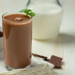 Szklanka kakao z mlekiem, ozdobiona miętą, obok dzbanek mleka i kawałki czekolady.