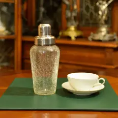 Szklany shaker do koktajli i biała filiżanka z podstawkiem na zielonej podkładce.