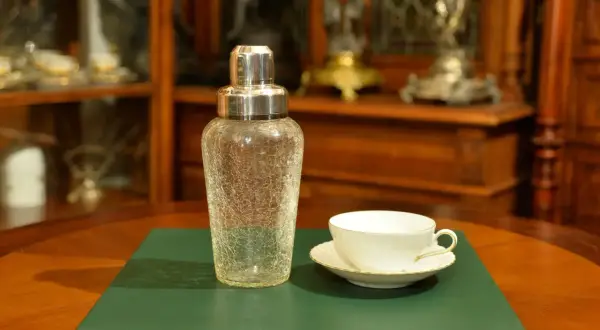 Szklany shaker do koktajli i biała filiżanka z podstawkiem na zielonej podkładce.