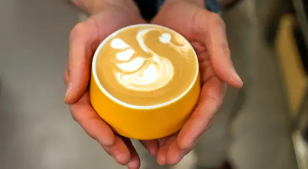 Dłonie trzymają żółtą filiżankę z latte art w kształcie łabędzia.