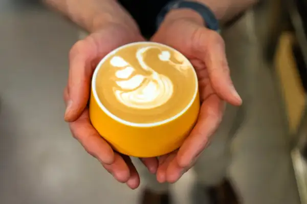 Dłonie trzymają żółtą filiżankę z latte art w kształcie łabędzia.